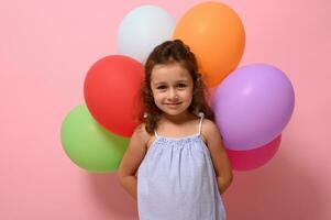 Confident portrait of gorgeous little birthday 4 years girl holding multicolored balloons behind back and smiling looking at camera posing against pink background with copy space photo