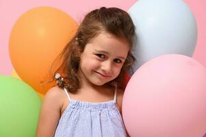 hermosa cumpleaños 4 4 años niña sonriente mirando a cámara, en pie detrás multicolor globos , aislado terminado rosado fondo, Copiar espacio. retrato de maravilloso pequeño niña para publicidad. foto