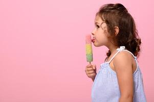 linda bebé niña disfrutando delicioso vistoso hielo crema paleta de hielo en palo aislado en rosado antecedentes con Copiar espacio. pequeño bebé comiendo vegano de colores sorbete foto