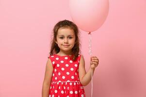 Birthday girl dressed in dress with polka-dots pattern holding pastel pink balloon, smiling, isolated over pink background with copy space. Close-up portrait of beautiful 4 years child for advertising photo