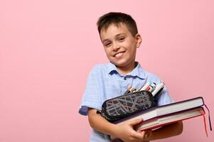 Smiling school boy posing over pink background with books and pencil case full of stationery. Back to school. Concepts with facial emotions and copy space for text photo