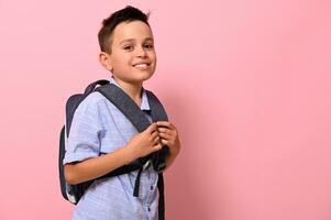 Side portrait of a smiling with toothy smile schoolboy with a school bag on his back on pink background with copy space. Back to school concepts photo