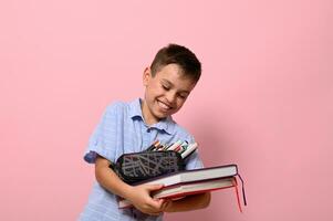 A school boy with pencil case and books laughing , posing over pink background with copy space. Concepts of back to school with facial expressions and emotions photo