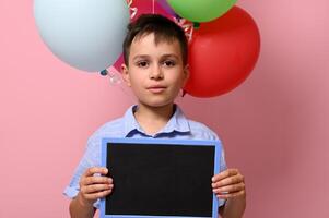 hermoso chico con blanco pizarra en su mano en pie en contra multicolor globos en rosado antecedentes con Copiar espacio foto