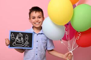 Cute boy holds multicolored colorful balloons in one hand and a chalkboard with drawn birthday cake in the other. Isolated over pink background with copy space photo