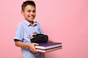 A smiling boy, student at school, holds books and pencil case in front of him. Back to school concepts on pink background with copy space photo