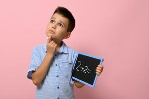 Cute schoolboy holding a chalkboard with chalk writing on it, thoughtfully solves a math problem. Isolated on pink background with copy space photo