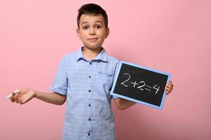 adorable chico, elemental colegio estudiante sostiene tiza y pizarra y resuelve matemáticas problema. rosado antecedentes espacio para texto. espalda a escuela. conceptos foto