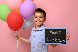 Happy teenage boy holds multicolored colorful balloons in one hand and a chalkboard with lettering Happy Birthday in the other. Isolated over pink background with copy space photo