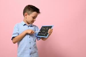 Lettering Back To School on a chalk board in the hands of a happy pupil. Isolated over pink background with copy space photo