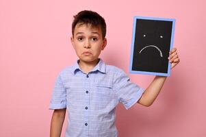 A frustrated boy in a blue shirt holds a blackboard near his face with a painted drawn expressing frustration, sadness and disappointment. Pink background, copy space photo