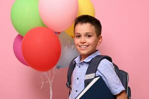 colegial con mochila participación libro y multicolor globos, linda sonriente posando a cámara terminado rosado antecedentes con Copiar espacio. conceptos de contento espalda a escuela. foto