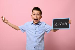 An adorable boy, an elementary school student, holds a chalk and a chalkboard and happily solves math problems. Pink background for text photo