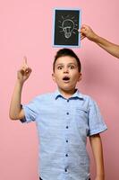 Chalkboard with drawn lamp, idea symbol, and schoolboy looking pensively with forefinger pointing on a pink background with copy space. Emotions on backgrounds photo