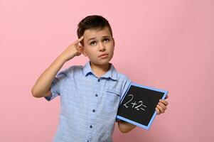 A schoolboy with a blackboard in his hand holds a finger to his temple and with a thoughtful look solves an arithmetic problem, standing on a pink background with a place for text. Back to school. photo