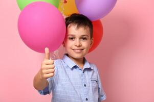 Handsome schoolboy showing thumb up to camera while posing against pink background with multicolored colorful balloons. Concepts photo