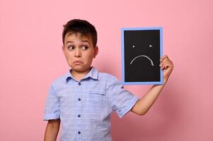 Isolated portrait on pink background of a sad schoolboy holding near his face a board with drawn sad emotion. Space for text photo