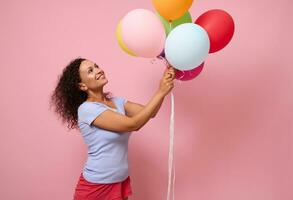 Winsome beautiful curly haired woman in blue t-shirt standing against pink colored background, smiling looking up at colorful bright air balloons in her hands. Anniversary concept with copy space photo