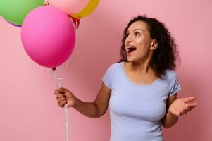 Cheerful African American young woman in blue t-shirt poses against a pink colored background with colorful bright inflated air balls in her hands looking at them with surprised facial expression. photo