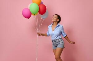 encantador sonriente joven mezclado carrera africano americano mujer con manojo de vistoso arco iris globos mirando arriba y teniendo divertido, vistiendo mezclilla pantalones cortos y azul camisa en rosado pared antecedentes con Copiar espacio foto