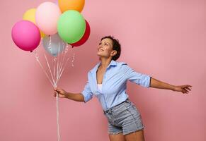 contento sonriente joven mezclado carrera africano americano mujer con manojo de vistoso arco iris globos mirando arriba y teniendo divertido, vistiendo mezclilla pantalones cortos y azul camisa en rosado pared antecedentes con Copiar espacio foto