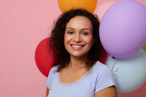 Close-up of attractive happy cheerful mixed race woman smiling with beautiful toothy smile looking at camera, posing against inflated colorful air balloons on pink colored background with copy space photo