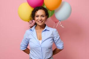 hermosa medio Envejecido mezclado carrera maravilloso mujer en azul camisa sonriente con con dientes sonrisa mirando a el cámara, participación multicolor aire pelotas detrás su atrás. rosado de colores antecedentes con Copiar espacio foto