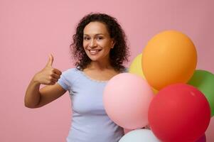 Attractive middle aged Hispanic curly haired woman in blue t-shirt showing a thumb up, smiling with toothy smile, holding multi-colored air balls posing against pink colored background with copy space photo