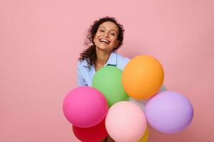 Close-up portrait of a charming joyful mixed race Hispanic woman with colorful multi-colored air balloons, laughing looking at camera, isolated on pink background with copy space. photo