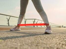 de cerca de el piernas de un atleta mujer, deportista en gris leggins haciendo cuerpo peso capacitación, hacer ejercicio con un resistencia aptitud elástico banda en un ciudad puente a amanecer foto