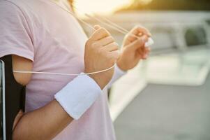 de cerca de hembra atleta manos con blanco terry pulseras participación auriculares en contra el amanecer cielo fondo, mientras hacer ejercicio al aire libre temprano en el Mañana. foto