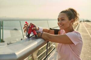 Confident relaxed middle aged sportswoman with a bottle of water and a skipping rope, admiring beautiful nature standing on the city bridge after intense cardio workout at sunrise photo