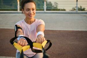 Close-up of beautiful middle aged Hispanic female athlete, sportswoman with earphones smiling cutely while exercising with suspension straps outdoor, enjoying body weight training early in the morning photo