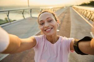 hermosa mujer, hembra atleta en rosado camiseta y pulseras sonrisas participación un teléfono inteligente en su extendido manos mientras haciendo un selfie durante Mañana empujoncito y rutina de ejercicio en el urbano puente a amanecer foto