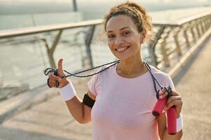 Delightful African female athlete with a jumping rope stands on the modern glass city bridge treadmill, smiles with a beautiful toothy smile looking at camera, enjoying morning cardio workout outdoor photo