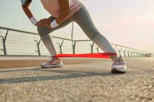 recortado imagen de un deportivo mujer, hembra atleta hacer ejercicio con elástico resistencia banda, extensión piernas, haciendo lado estocadas mientras ejecutando un peso corporal formación al aire libre en el ciudad puente a amanecer foto