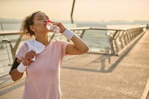 de edad mediana hembra atleta en un rosado camiseta y gris polainas, con blanco pulseras, sostiene un terry toalla en su espalda y bebidas agua después rutina de ejercicio en pie en el ciudad puente a amanecer foto