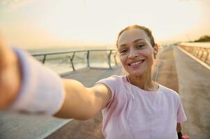 deportista, hembra atleta en rosado camiseta y pulseras sonrisas participación un móvil teléfono en su extendido manos mientras haciendo Auto retrato durante Mañana empujoncito y rutina de ejercicio en el puente a amanecer foto