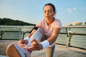 Female athlete with earphones, runner tying sneakers laces while enjoying morning jog on a city bridge treadmill. Run, jogging, healthy active lifestyle, cardio workout and outdoor fitness concept photo