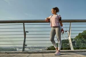 Full length of a delightful sportswoman, female athlete with a skipping rope, standing on the modern glass city bridge, relaxing after cardio workout outdoor. Sport, fitness, active lifestyle concept photo