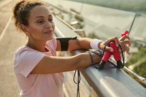 Confident relaxed middle aged sportswoman with a bottle of water and a skipping rope, looking away while admiring beautiful nature standing on the city bridge after intense cardio workout at sunrise photo