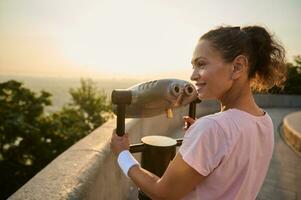 deportivo bonito mujer mirando mediante un telescopio a el ciudad. atlético mujer en un rosado camiseta es acecho el hermosa verde naturaleza y el río antecedentes mediante estacionario ciudad prismáticos foto