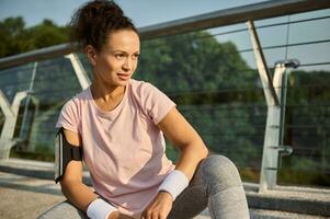 Attractive determined African American female athlete, beautiful sportswoman in pink t-shirt looking away sitting on a squat position on the glass city bridge, enjoying sunny morning workout photo