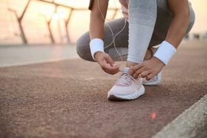 Cropped image of a fitness woman tying shoelaces on sneakers and getting ready for morning running and sports workout outdoors on a city bridge treadmill at sunrise photo