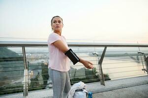 Middle-aged athlete in pink t-shirt puts on a smartphone holder on her hand, getting ready for a morning run, stands on a treadmill on a pedestrian city bridge and looking aside. Sport, motivation photo