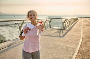 un de edad mediana deportista en un rosado camiseta y gris polainas, con blanco pulseras, sostiene un terry toalla en su espalda y sonrisas alegremente, demostración un botella de agua a el cámara foto