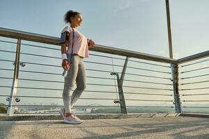 Full length portrait of a sportswoman, female athlete with a skipping rope, standing on the modern glass city bridge, relaxing after cardio workout outdoor. Sport, fitness, active lifestyle concept photo