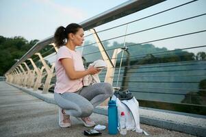 Confident athletic middle-aged woman in pink t-shirt and sports leggings sits on bridge and unwraps clothes, holds terry white towel, prepares for morning run and outdoor workout on early summer day photo
