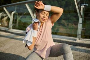 Beautiful delighted sportswoman wiping her face and neck from sweat with white terry towel and wristband, sitting on squat position on the bridge, relaxing after jog on a sunny summer day photo