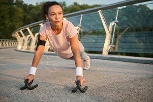 medio adulto Rizado peludo deportista en rosado camiseta y gris leggins con teléfono inteligente poseedor y pulseras, haciendo Lagartijas al aire libre en el ciudad puente. físico ejercicio y sano estilo de vida concepto. foto